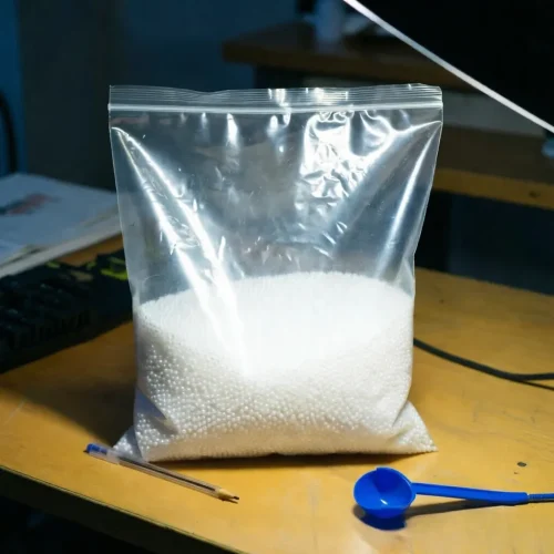 eva-resin-pellet-sample-in-zip-lock-bag A small zip-lock bag filled with white EVA resin pellets as a lab sample, placed on a desk with a blue scoop and pen, highlighting the fine quality of the raw plastic material.