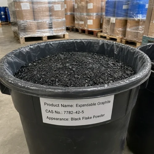 Open black plastic drum filled with black flake powder of Expandable Graphite CAS 7782-42-5, labeled with appearance description, in industrial warehouse with stacked drums in background.