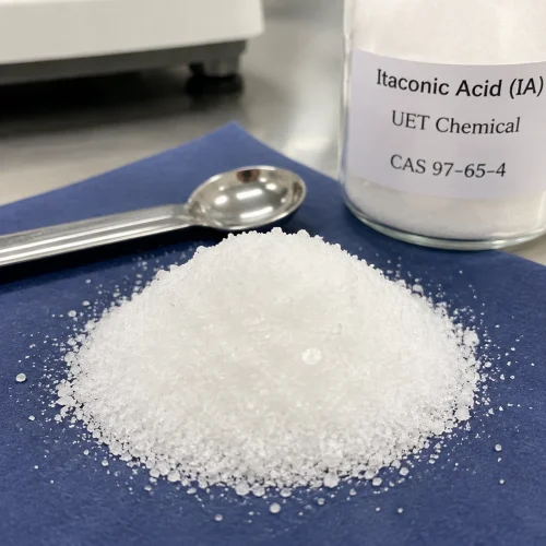 White crystalline Itaconic Acid (IA) granules (CAS 97-65-4) on a blue mat beside a steel spoon and UET Chemical sample bottle