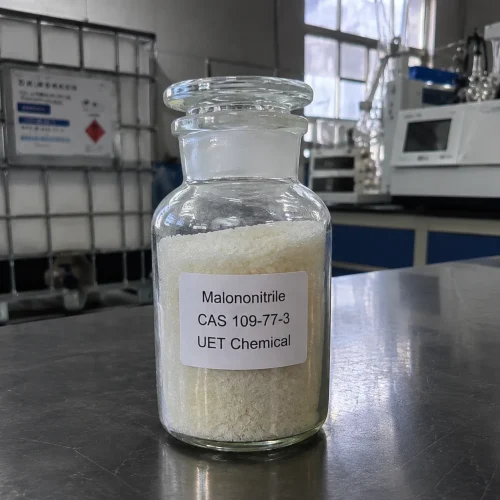 malononitrile-cas-109-77-3-glass-sample-bottle-crystalline-solid Glass reagent bottle with ground glass stopper containing white crystalline solid Malononitrile (CAS 109-77-3) on stainless steel lab bench, labeled UET Chemical, IBC tank visible in background