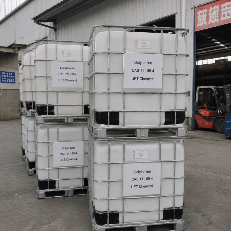 n-octylamine-cas-111-86-4-ibc-tote-bulk-packaging Stacked white IBC totes with metal cages containing Octylamine (CAS 111-86-4), labeled UET Chemical, in outdoor loading area with forklift in background
