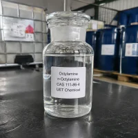 octylamine-n-octylamine-cas-111-86-4-glass-sample-bottle Glass reagent bottle with ground glass stopper containing clear liquid Octylamine (n-Octylamine, CAS 111-86-4) on dark tabletop, labeled UET Chemical, with IBC and blue drums in background
