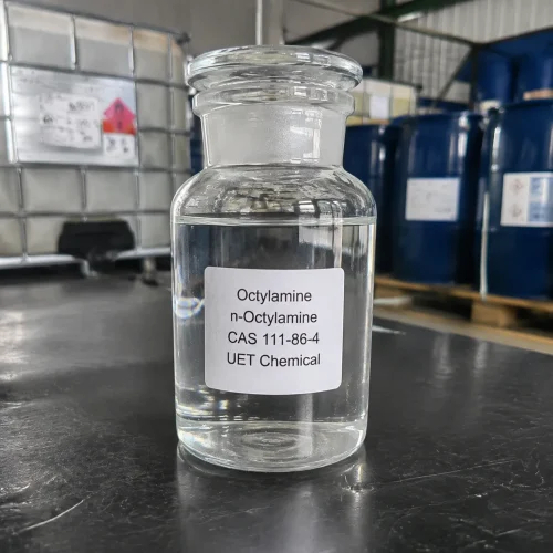 octylamine-n-octylamine-cas-111-86-4-glass-sample-bottle Glass reagent bottle with ground glass stopper containing clear liquid Octylamine (n-Octylamine, CAS 111-86-4) on dark tabletop, labeled UET Chemical, with IBC and blue drums in background