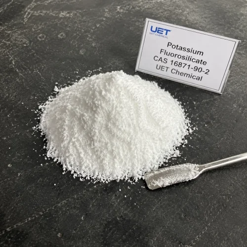 Pile of fine white crystalline powder Potassium Fluorosilicate (CAS 16871-90-2) on dark stone surface with metal spatula, labeled UET Chemical, sample for glass, ceramic or enamel industry raw material verification