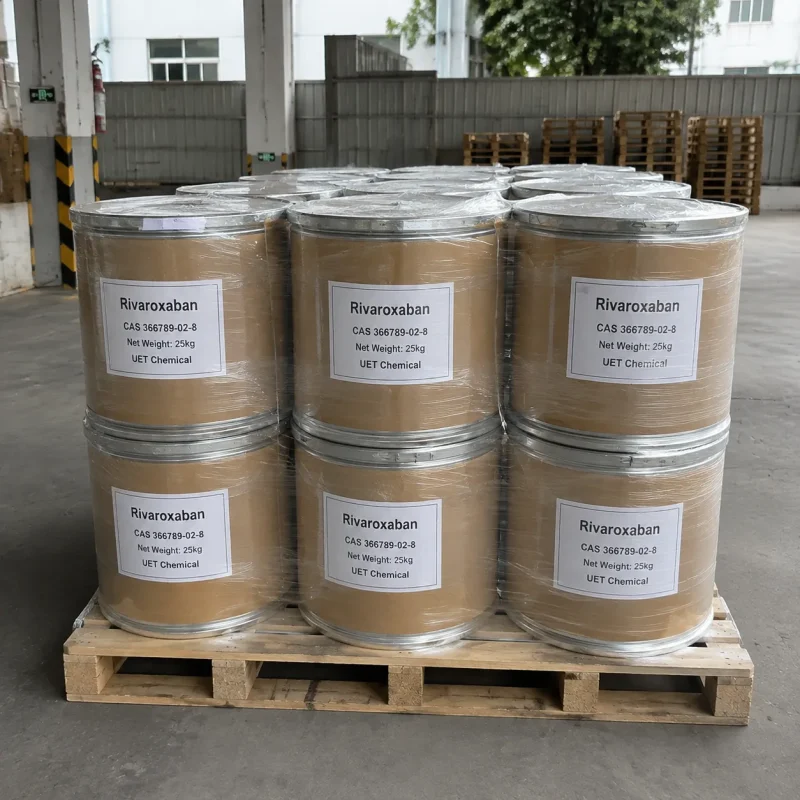 rivaroxaban-cas-366789-02-8-25kg-fiber-drum-pallet Stacked 25kg brown fiber drums of Rivaroxaban CAS 366789-02-8 on wooden pallet, wrapped in plastic film for protection, inside a warehouse with safety signage and spare pallets visible.