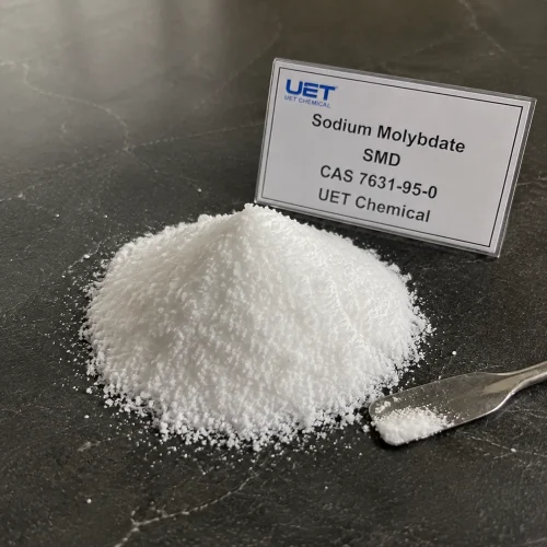 Pile of fine white crystalline powder Sodium Molybdate (CAS 7631-95-0) on dark stone surface with metal spatula, labeled UET Chemical, sample for fertilizer additive or trace element verification