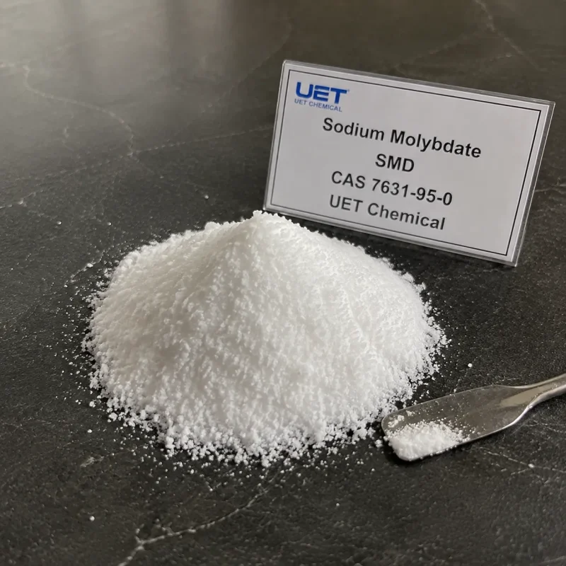sodium-molybdate-cas-7631-95-0-white-powder-sample Pile of fine white crystalline powder Sodium Molybdate (CAS 7631-95-0) on dark stone surface with metal spatula, labeled UET Chemical, sample for fertilizer additive or trace element verification