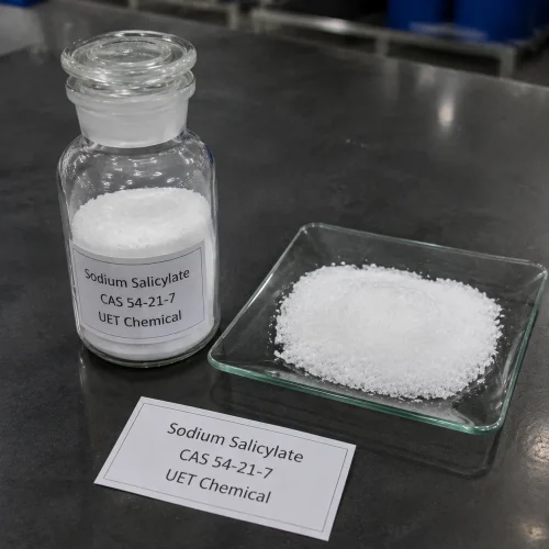 sodium-salicylate-white-powder-sample-glass-bottle Close-up of white crystalline powder Sodium Salicylate (CAS 54-21-7) in a clear glass reagent bottle with ground glass stopper, alongside a glass dish displaying the powder texture, labeled UET Chemical, product sample on a dark lab bench