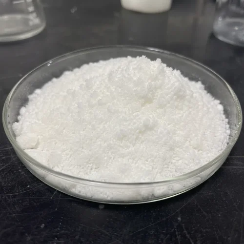 White crystalline Sodium Stannate Trihydrate CAS 12209-98-2 sample in a glass petri dish, on a lab bench with glassware in the background.