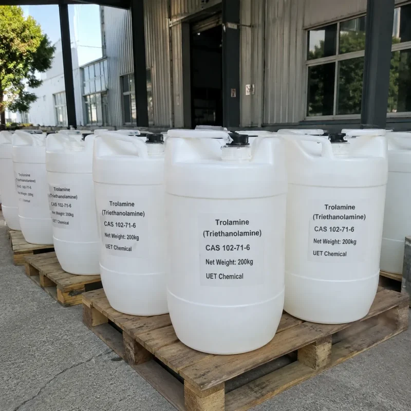 Multiple 200kg white plastic drums of Trolamine (Triethanolamine) CAS 102-71-6 stacked on wooden pallets outdoors, with factory building and greenery in background.