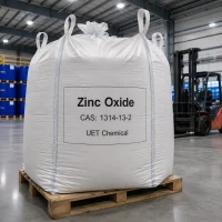 Large white jumbo bag (ton bag) of Zinc Oxide CAS 1314-13-2 on a black plastic pallet in a warehouse, label showing UET Chemical branding, with IBC tanks visible in the background.