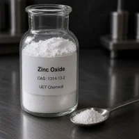 Fine white powder sample of Zinc Oxide CAS 1314-13-2 in a transparent glass reagent bottle with a stainless steel spoon beside it on a dark metal lab bench, label showing UET Chemical branding.